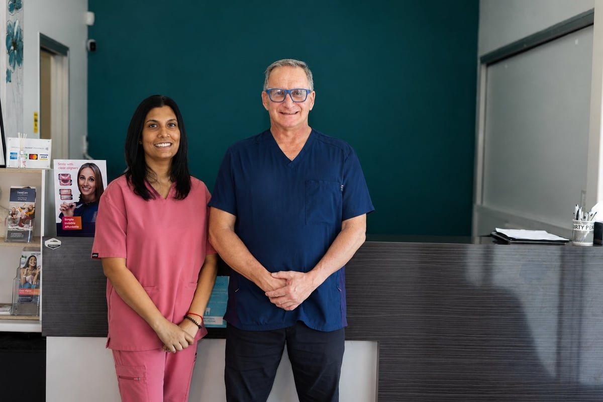 Two people standing in a medical office, woman in pink scrubs and man in navy shirt and glasses