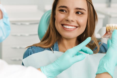 Young woman smiling at dental office wearing denim shirt with dental professionals gloved hands holding mirror and tools nearby