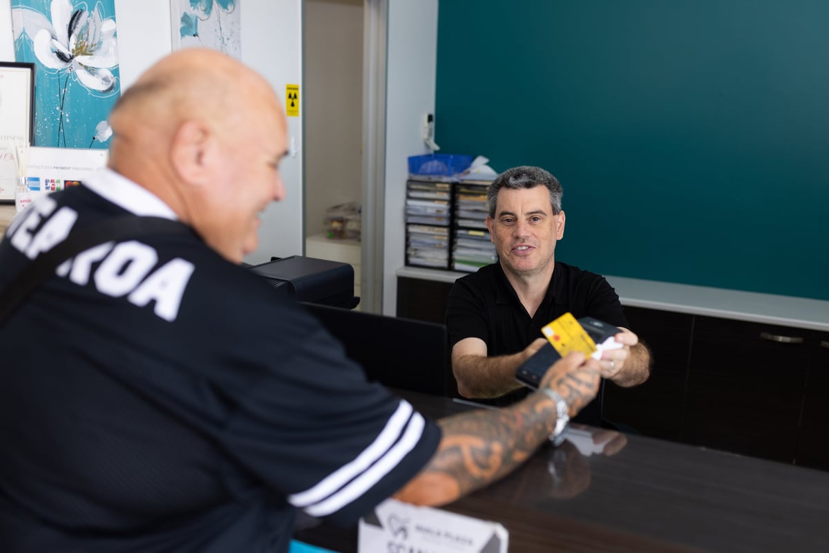 Two men in a gym setting, one wearing a black shirt receiving first aid or medical treatment on his arm while the other man applies a yellow bandage or wrap.