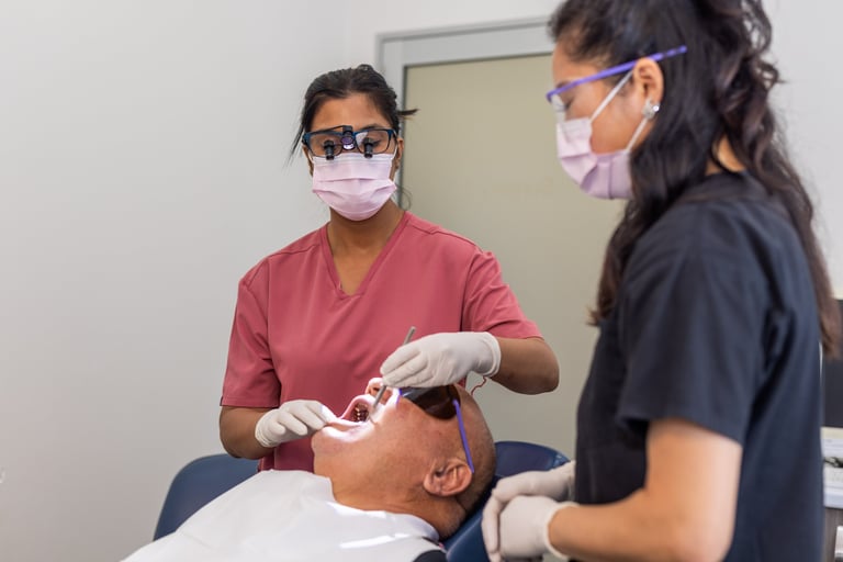 Two dental professionals wearing masks and protective eyewear examine a patients teeth during a dental procedure in an office setting.